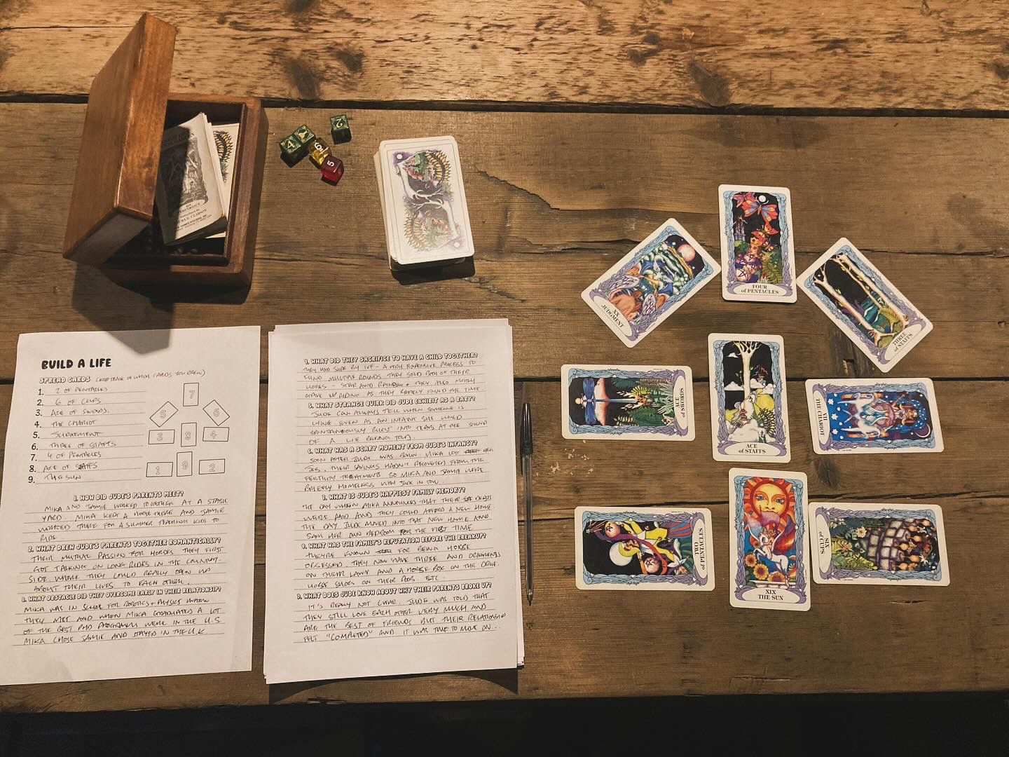 A nine card tarot spread on a wooden table next to a couple of scrawled pages of workbook answers. A complete Build a Life spread from the upcoming game, Jude's World