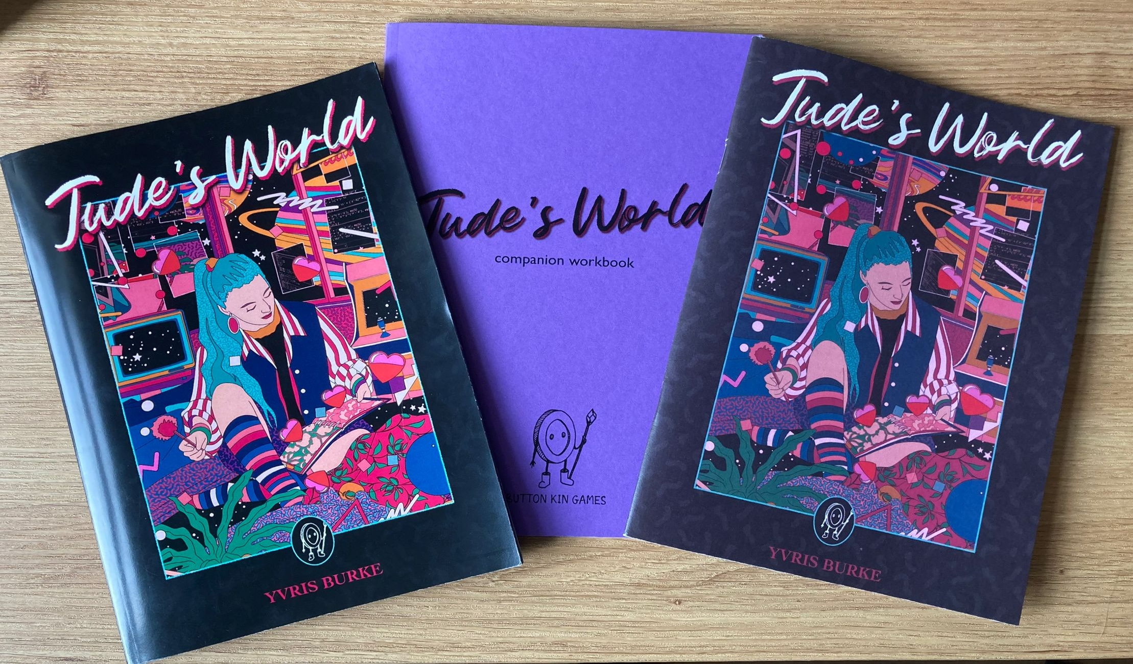 Three books on a wooden desk. Two are Jude's World zines with a colourful cover depicting a young person with turquoise hair writing in their diary. One is shiny, the other is matte. In the middle and slightly obscured by the zines is a purple workbook with Jude's World companion workbook on the cover